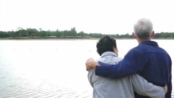 Видео счастливой азиатской пожилой пары, разговаривающей и смотрящей на озеро с горным фоном — стоковое видео