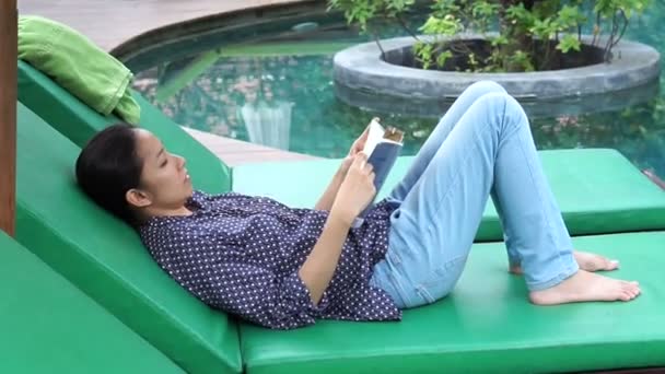 Mulher asiática senta-se ao lado da piscina e livro de leitura — Vídeo de Stock