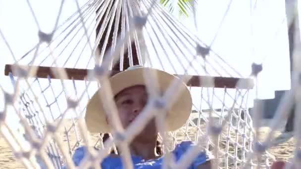 Видео Азиатка, девушка в шляпе расслабиться на гамаке на солнечном пляже с кокосовыми пальмами — стоковое видео