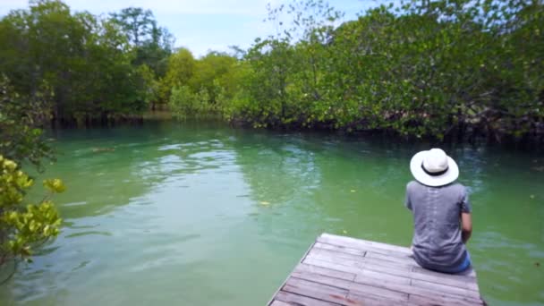 Video de chica asiática con sombrero sentado en un pequeño muelle al lado del bosque intermareal — Vídeo de stock