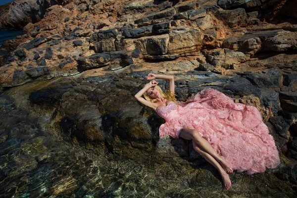 Hermosa mujer rubia sexy vestido de salón rosa de pie sobre las rocas en Santorini — Foto de Stock