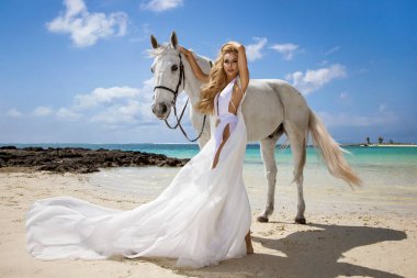 Akan beyaz elbiseli zarif bir kadın tropikal plajda görkemli beyaz bir atla poz veriyor, lüks moda fotoşopu, egzotik deniz kıyısı cazibesi ve Mauritius adası manzarasında güzellik.