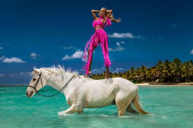 Turkuaz okyanus gölünde beyaz bir atın üzerinde duran canlı pembe püsküllü elbiseler içinde manken Mauritius tropikal moda fotoshoot