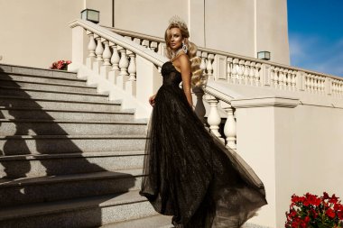 Parlak siyah balo elbisesi ve elmas tacıyla şık bir kadın lüks saray merdivenlerinde duruyor.