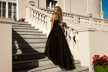 Parlak siyah balo elbisesi ve elmas tacıyla şık bir kadın lüks saray merdivenlerinde duruyor.