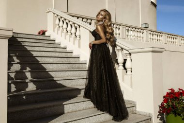 Parlak siyah balo elbisesi ve elmas tacıyla şık bir kadın lüks saray merdivenlerinde duruyor.