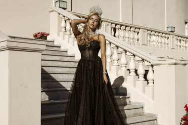 Parlak siyah balo elbisesi ve elmas tacıyla şık bir kadın lüks saray merdivenlerinde duruyor.