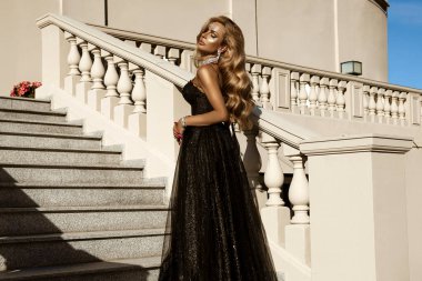 Parlak siyah balo elbisesi ve elmas tacıyla şık bir kadın lüks saray merdivenlerinde duruyor.