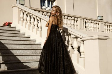 Parlak siyah balo elbisesi ve elmas tacıyla şık bir kadın lüks saray merdivenlerinde duruyor.