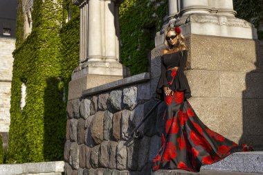 Flamenko 'dan esinlenilmiş siyah ve kırmızı gül desenli elbisesiyle tarihi taş duvara poz veren kadın.