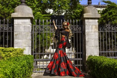 Güneşli bir günde, saray bahçesinde metal bir kapının önünde duran zarif siyah ve kırmızı çiçekli bir elbise içinde romantik bir kadın.