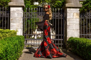 Güneşli bir günde, saray bahçesinde metal bir kapının önünde duran zarif siyah ve kırmızı çiçekli bir elbise içinde romantik bir kadın.