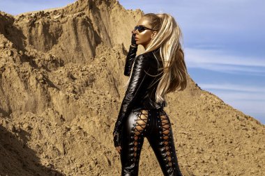 Siyah deri kıyafetli fütürist seksi model, dinamik hareketli çöl kum manzarasında güven içinde poz veriyor.