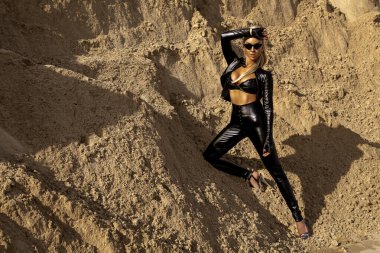 Siyah deri kıyafetli fütürist seksi model, dinamik hareketli çöl kum manzarasında güven içinde poz veriyor.