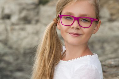 Young beautiful child girl model long curly blond hair smiling in pink glasses and a chic elegant dress at the rocks and the sea in Spain, Greece, Santorini