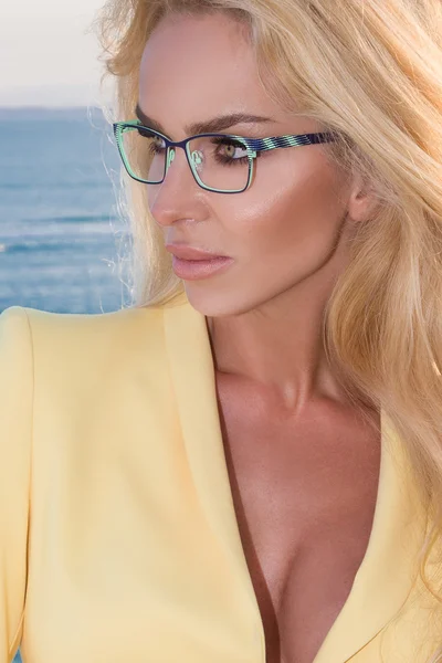 Beautiful blond hair sexy woman young girl model in blue and black color glasses in yellow elegant jacket, suit around the pool with a balustrade overlooking the sea and the island of Santorini