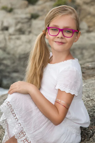Joven hermosa niña modelo largo pelo rubio rizado sonriendo en gafas de color rosa y un elegante vestido elegante en las rocas y el mar en España, Grecia, Santorini — Foto de Stock