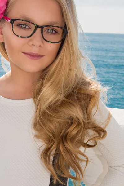 Young beautiful child girl model long curly blond hair smiling in pink glasses and a chic elegant long blacj and white dress at the pool and the sea in Spain, Greece, Santorini — стокове фото