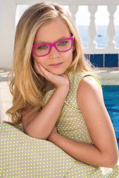 Unga vackra barn flicka modell långt lockigt blont hår leende i rosa glas och en chic grön lång klänning i rosa glas vid poolen med räcke och havet i Spanien, Grekland, Santorini — Stockfoto