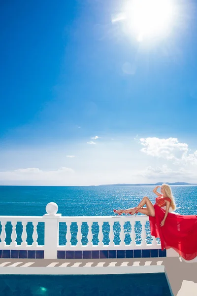 Симпатичні блондин сексуальна жінка модель дівчина басейн в червоній елегантні довгі сукні з довгий поїзд з кристалів тлом синього моря і білий балюстраді у Санторіні — стокове фото