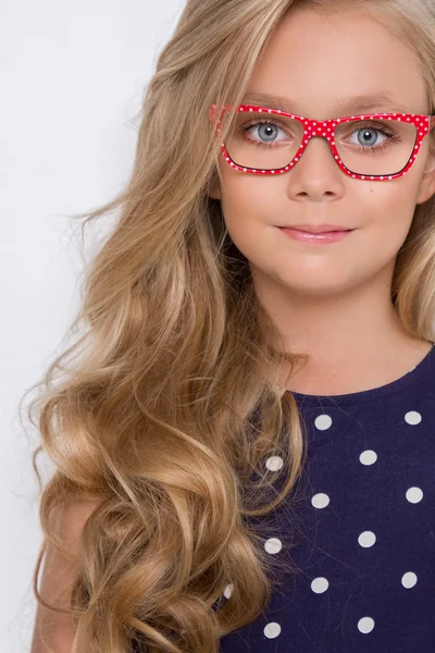 Retrato de una encantadora hija niña en pelo largo y rubio violeta, vestido azul con puntos blancos y gafas rojas con puntos blancos mira a la cámara, foto en el fondo blanco ojos increíbles — Foto de Stock