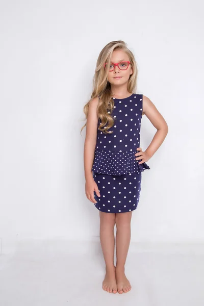 Portrait of a lovely little girl daughter in long blond hair and violet, blue dress with white dots and red glasses with white dots looks at the camera, photo on the white background amazing eyes — Stockfoto