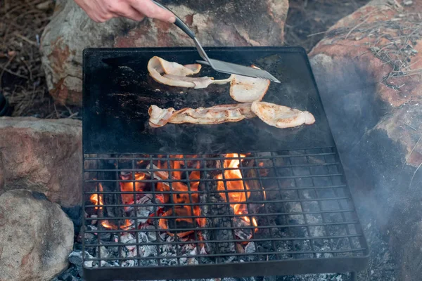 Breakfast camp cooking. Grilling crispy bacon on a cast iron plate over the camp fire. Camping lifesyle