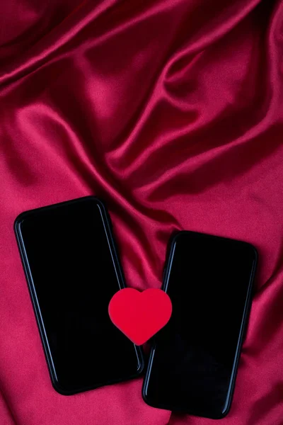 Red heart and two mobile phones on red silk fabric with waves. Internet dating, copy space, Valentines day concept.