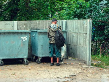 Kirli giysiler içinde aç ve fakir evsiz bir adam şehirdeki çöp konteynırında yiyecek arıyor. Modern toplumun sosyal sorunları. Sokak serserilerinin yaşam tarzı, sokaklarda yaşamak..