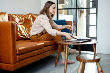 Evdeki kanepede dizüstü bilgisayarlı bir kadın oturuyor.