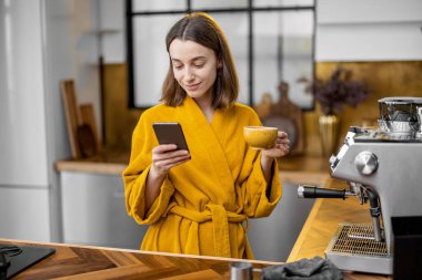 Sabahları mutfakta kahve ve telefon taşıyan bir kadın.