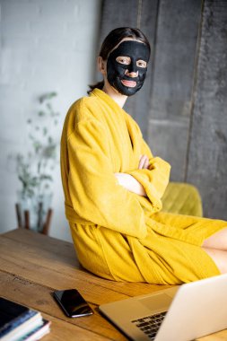 Ofiste yüzünde siyah temizlik maskesi olan sarı bornozlu bir kadın.