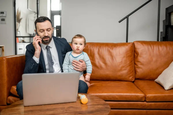 Mladý táta pracuje na notebooku a mluví po telefonu — Stock fotografie