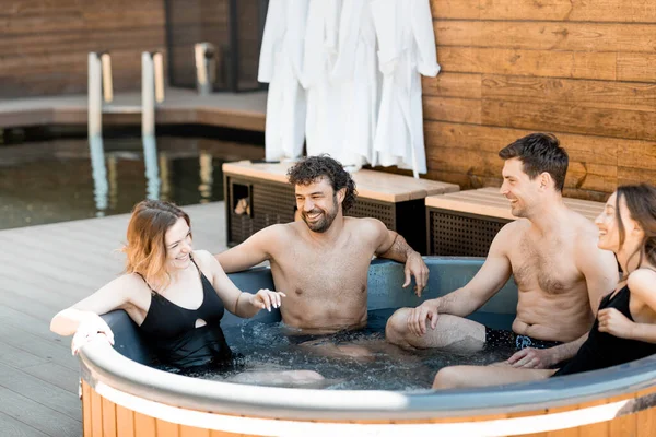 Compañía de amigos que humean en una bañera al aire libre cerca del spa y el lago —  Fotos de Stock