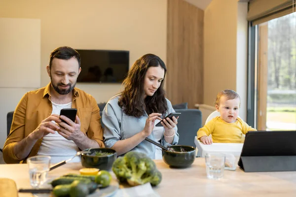 Rodiče a jejich roční chlapeček sedí v mobilních zařízeních během oběda doma — Stock fotografie