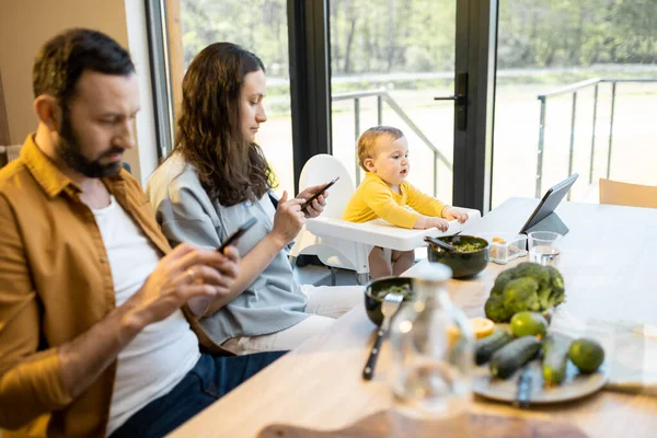 Vanhemmat ja heidän yhden vuoden vauva poika istuu mobiililaitteissa lounaan aikana kotona — kuvapankkivalokuva