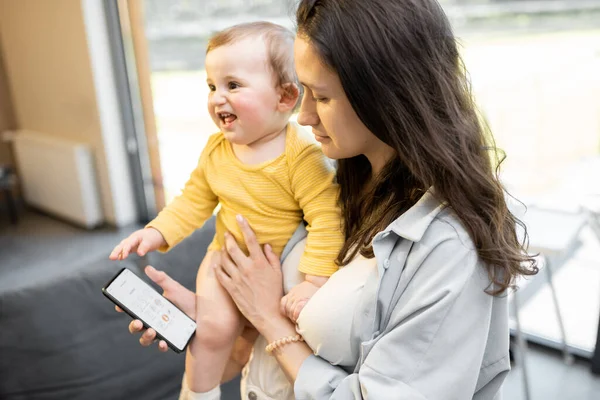 Mamma styra hem enheter med smartphone, står med ett barn hemma — Stockfoto