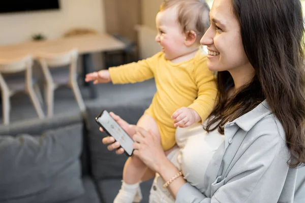 Mamma kontrollerer hjemmeutstyr med smarttelefon, står med en baby hjemme – stockfoto