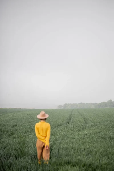 Vrouw met hoed loopt op groen tarweveld tijdens mistig weer — Stockfoto