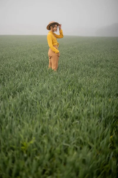 Žena v klobouku chůzi na zeleném pšeničném poli během mlhavého počasí — Stock fotografie