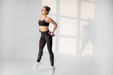 Beyaz odada halterli bir kadın kardiyo yapıyor.