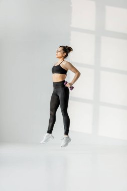 Beyaz odada halterli bir kadın kardiyo yapıyor.