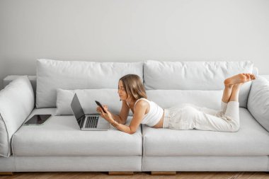 Genç kadın rahat koltukta telefon ve dizüstü bilgisayarla rahatça uzanıyor.
