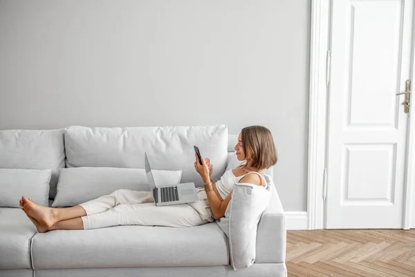 Mladá žena ležící uvolněná s telefonem a notebookem na pohodlném gauči — Stock fotografie