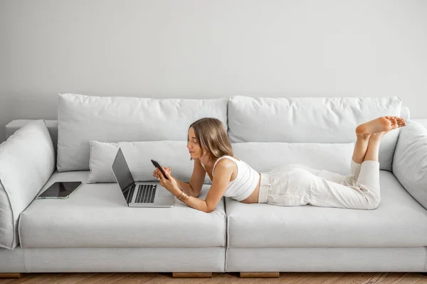Ung kvinna ligger avslappnad med telefon och laptop på den bekväma soffan — Stockfoto