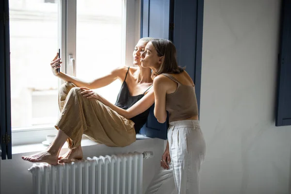 Dos mujeres se divierten, hablando en línea en el teléfono celular por la ventana — Foto de Stock