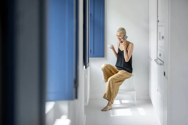 Nainen puhelimen ja kahvin kanssa lähellä ikkunaa kotona — kuvapankkivalokuva