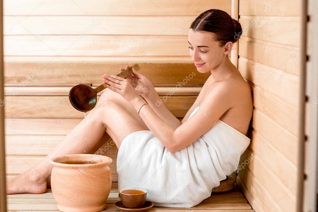 Young woman in white towel pouring water while resting in Finnish sauna.