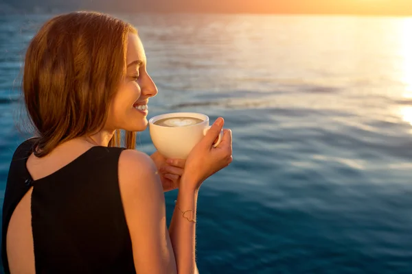Young woman drinking coffee on the pier at sunrise — Stock Photo, Image