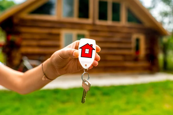 Holding house key with wooden cottage on background — Stock Photo, Image